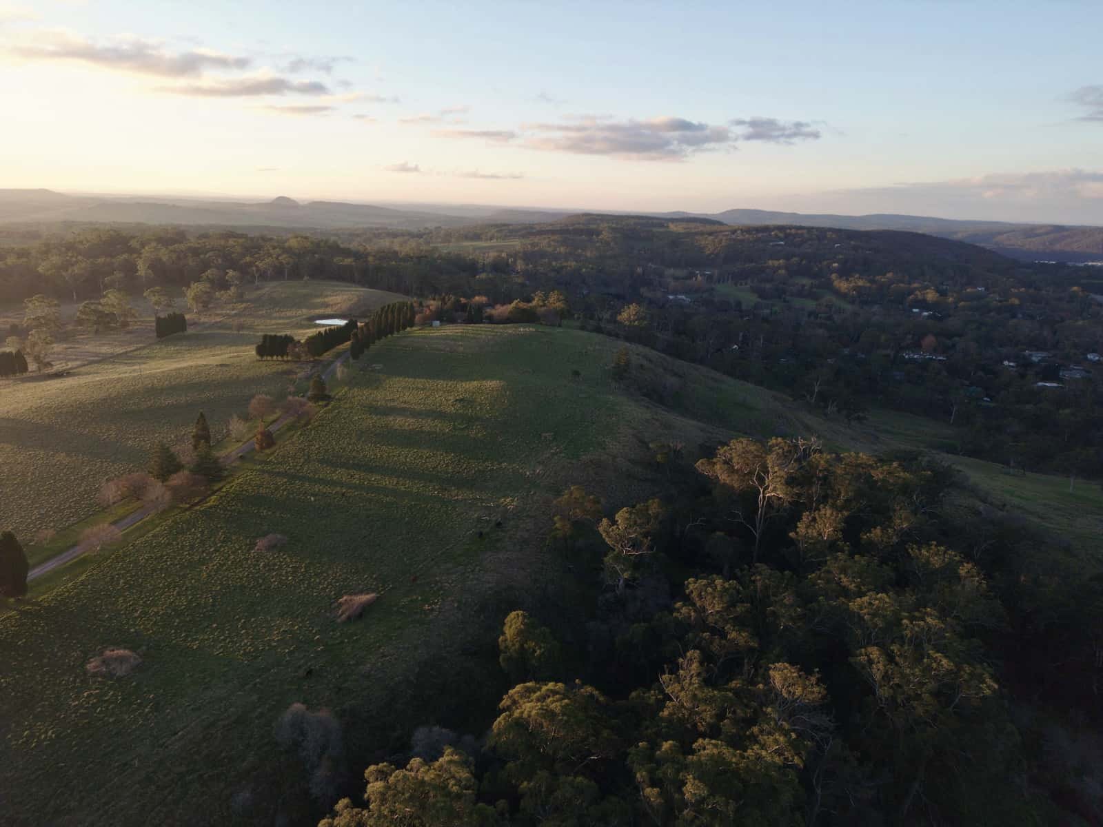 Green hills and paddocks roll away under a hazy evening sky, with stands of trees along the ridges.
